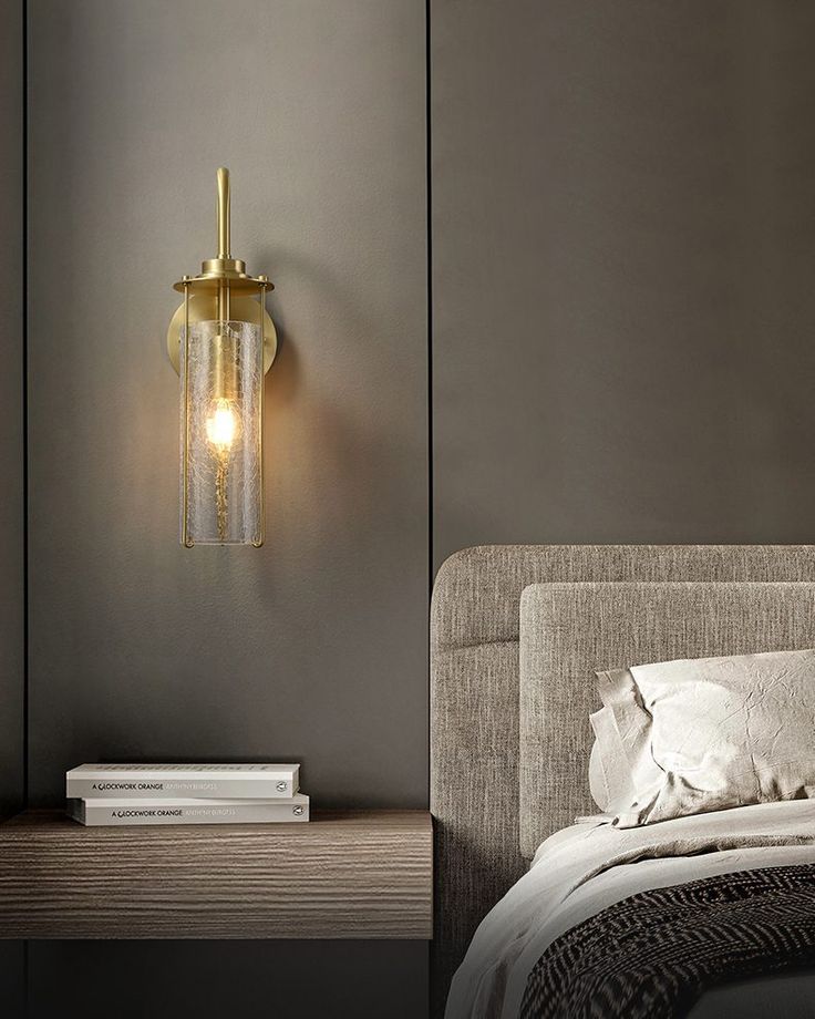 Gold wall sconce on a gray wall above a wooden nightstand with books, next to a bed with gray bedding.