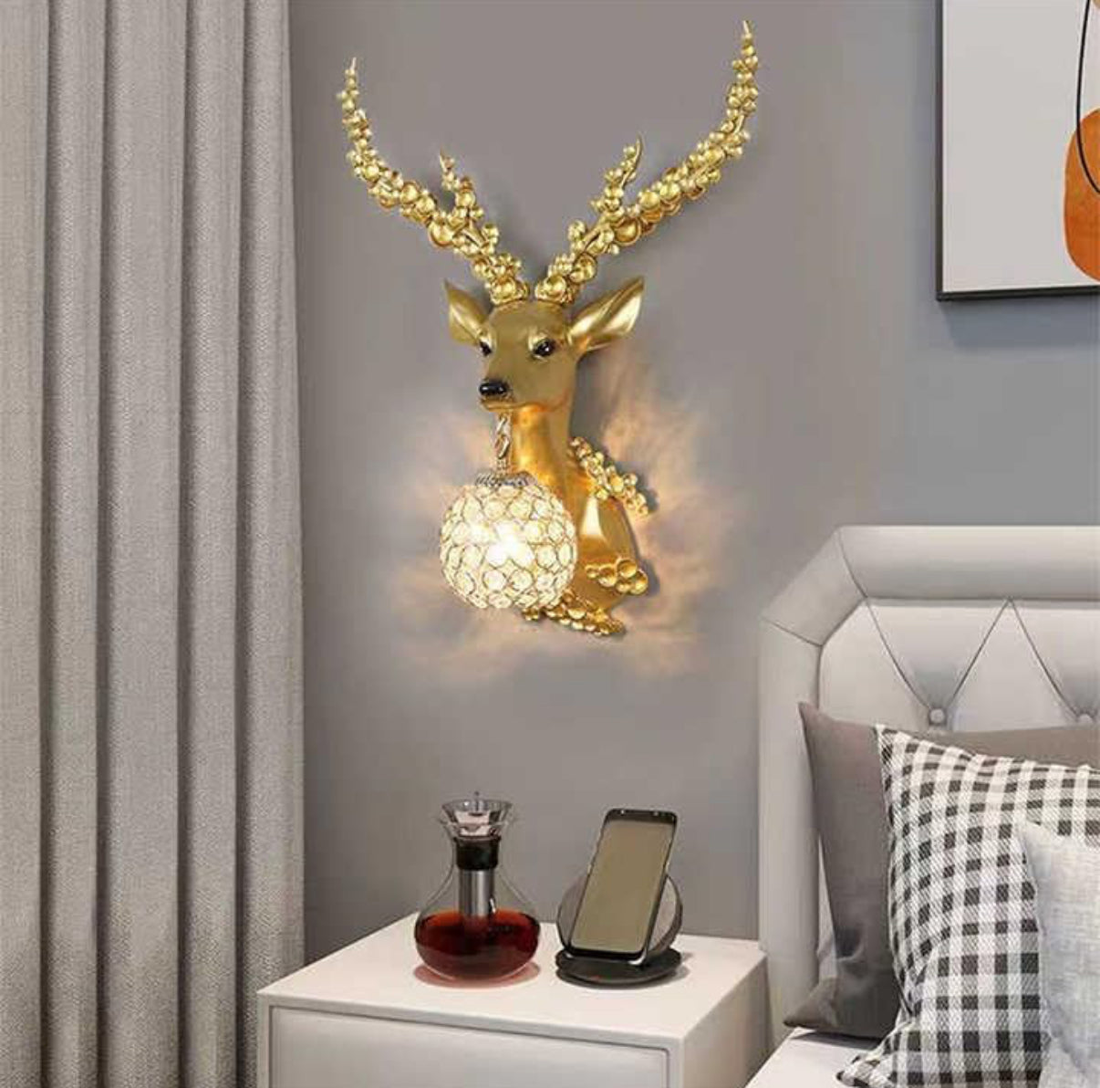 Decorative wall light shaped like a deer head on a wall above a nightstand with a phone and bottle.