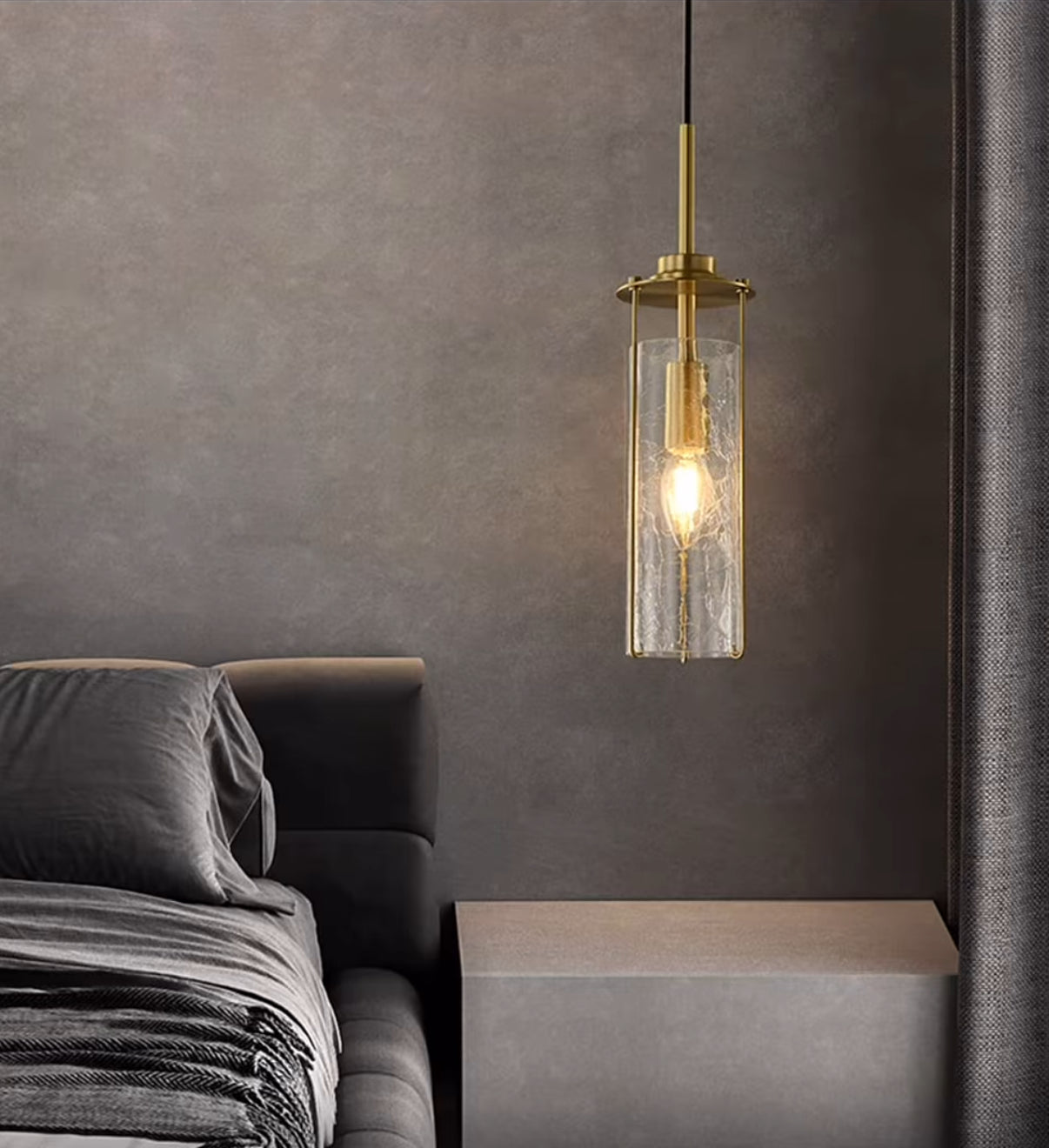 Gold pendant light with clear glass shade hanging above a bed in a bedroom.