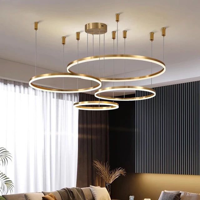 A modern white LED chandelier with gold accents, featuring four concentric rings, hanging in a room with a dining area and a contemporary sofa.
